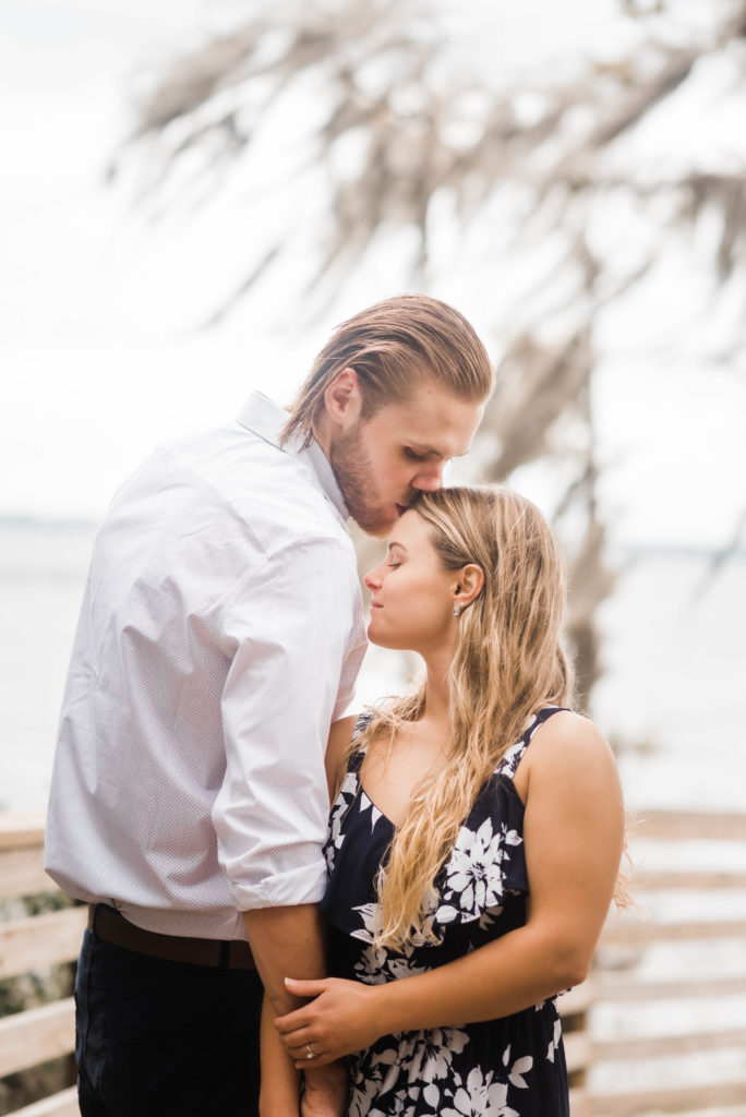 Groom kisses bride on forehead while taking engagement photo the dock of the St Johns River at the   Alpine Groves Park. in Jacksonville Florida  Photo By Black Tie & Co. www.btcweddings.com